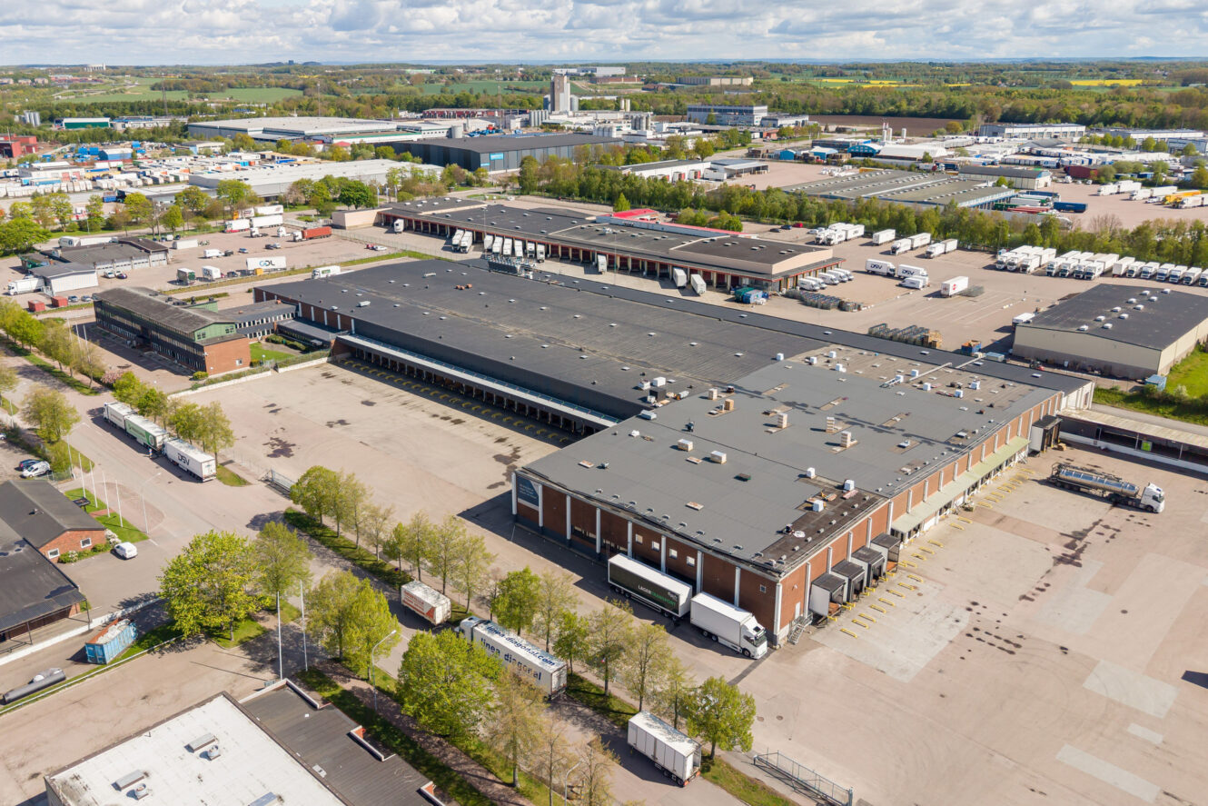 Flygbild över ett stort industriellt lagerkomplex med flera lastkajer, lastbilar och omgivande anläggningar i ett industriområde.