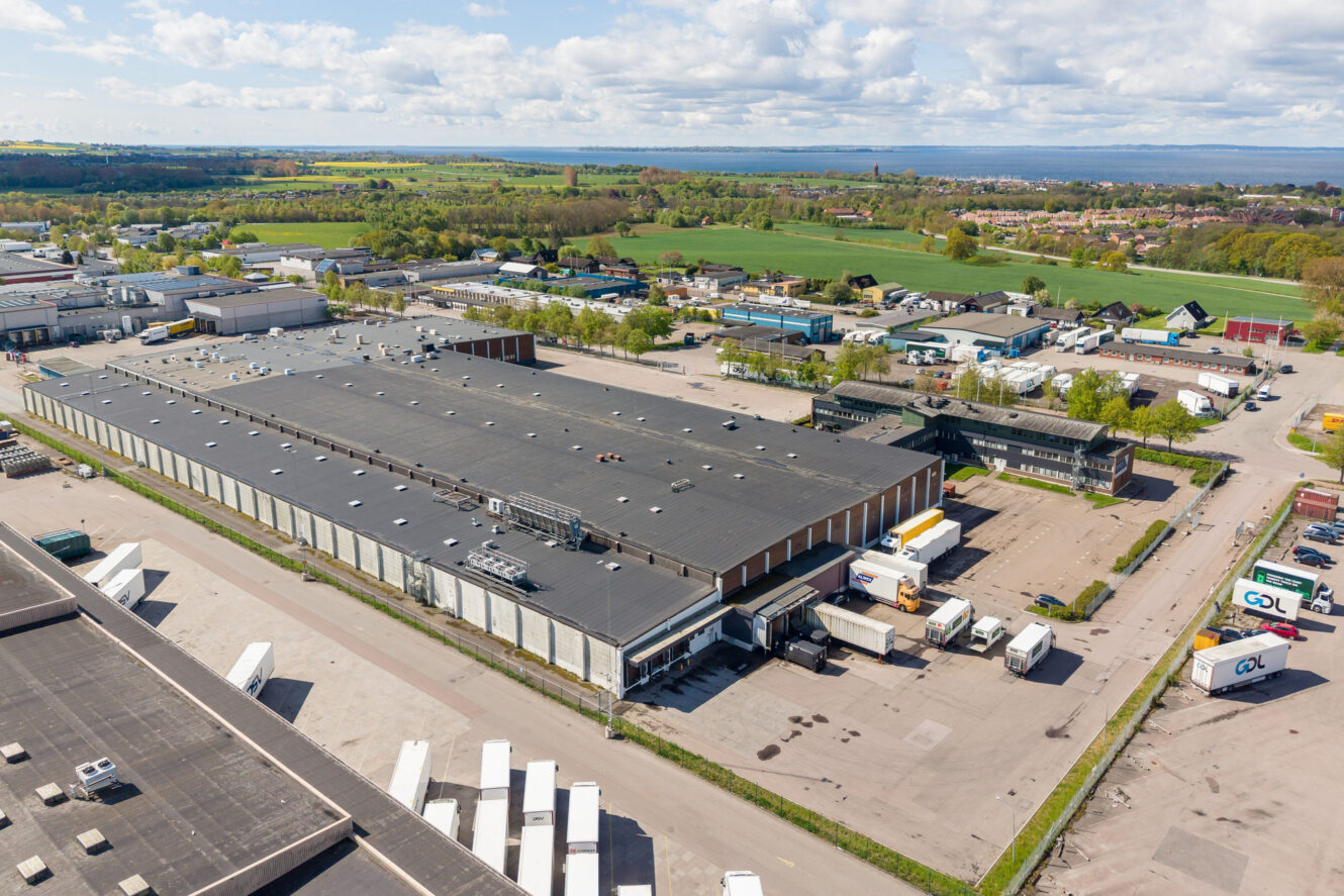Flygbild över ett stort lager med lastkajer, lastbilar parkerade utanför, industribyggnader och gröna fält i bakgrunden.