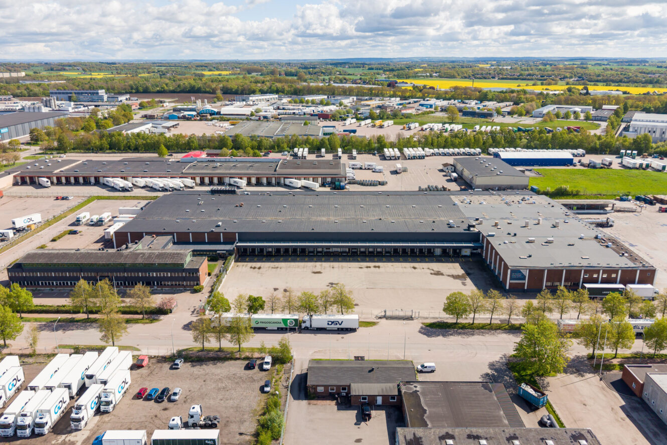 Flygbild över ett vidsträckt industriellt lagerkomplex med lastkajer, lastbilar och närliggande byggnader i ett halvlantligt landskap.