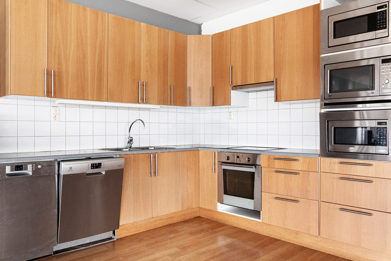 Modernt kök med träskåp, vitvaror i rostfritt stål inklusive två diskmaskiner, en ugn och tre staplade mikrovågsugnar, vitkaklat bakplank och trägolv.