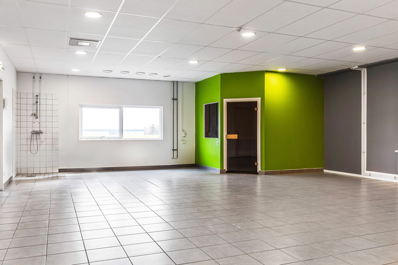 Ett rymligt, tomt rum med grått klinkergolv, vitt tak, en grön hörnstruktur med fönster och dörr samt ett kaklat väggparti med en duscharmatur.