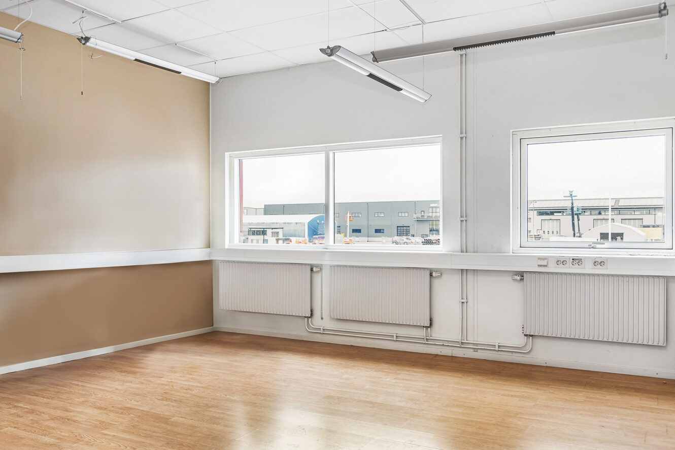 Tomt kontorsrum med ljust trägolv, beige och vita väggar, stora fönster och taklampor; industribyggnader syns utanför.