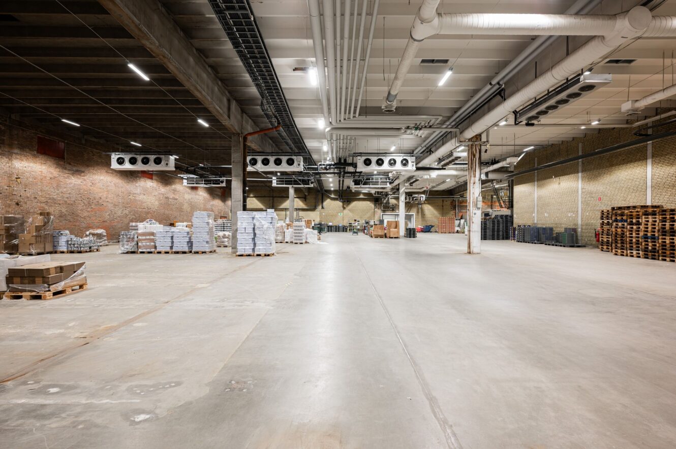 Stor, mestadels tom lagerlokal med betonggolv, synliga takventiler, staplade lastpallar och utspridda lådor längs väggarna.