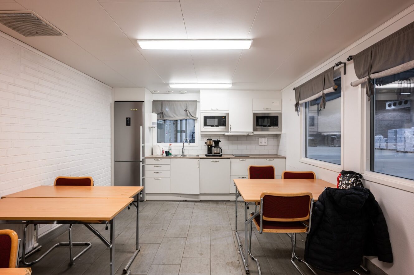 Ett litet, rent pentry med vita skåp, kylskåp, mikrovågsugn, kaffebryggare och två bord med stolar. En kappa och en väska är draperade över en stol nära fönstret.