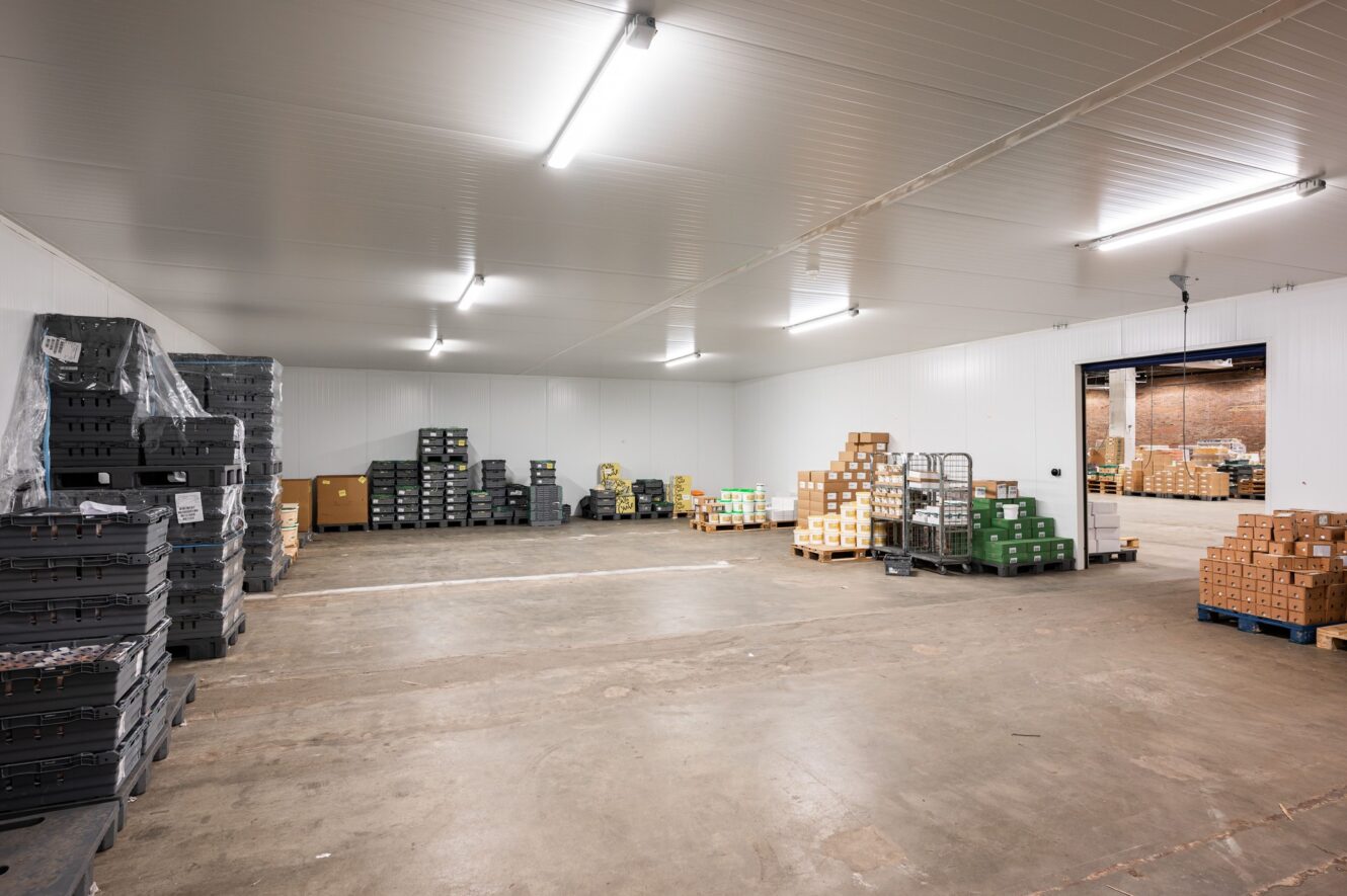 Ett stort lagerrum med staplade plastbackar och lådor på pallar, lysrörsbelysning och en öppen lastbryggedörr.