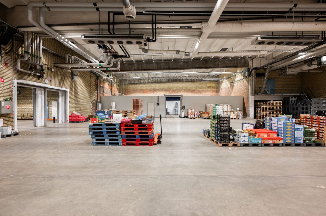 Stor lagerlokal med synliga rör och pallar staplade med olika lådor och backar, organiserade i öppna utrymmen på betonggolvet.