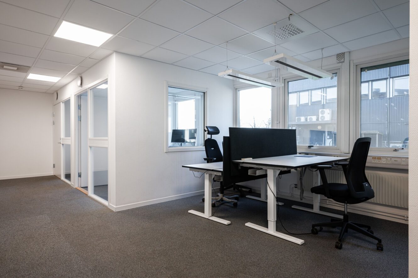 Modernt kontorslandskap med två tomma skrivbord och stolar vid stora fönster, vita väggar och mötesrum med glasväggar i bakgrunden.