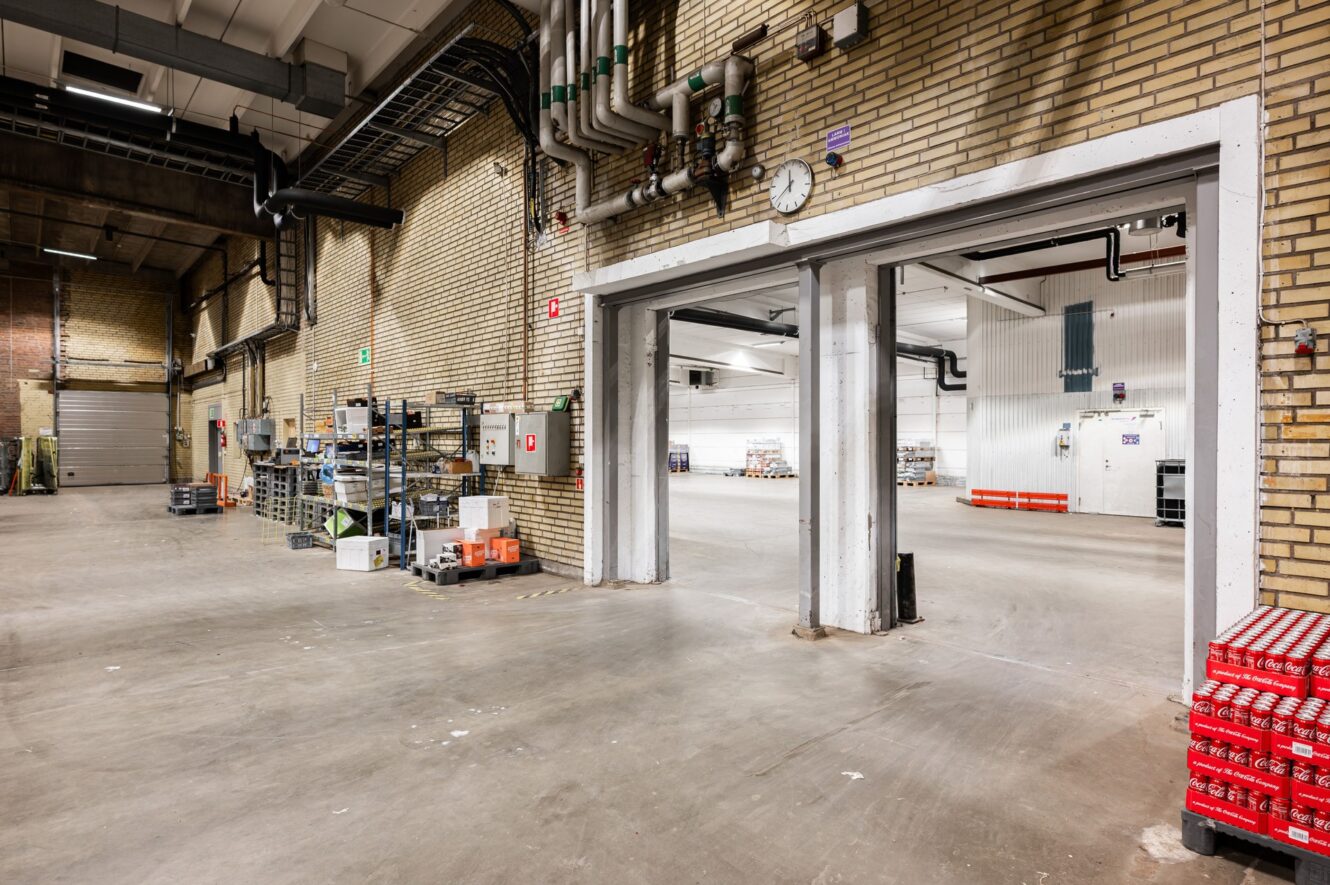 En rymlig industriell lagerlokal med exponerade tegelväggar, hyllor, synliga rör och öppna dörrar som leder till ett starkt upplyst lagerutrymme.