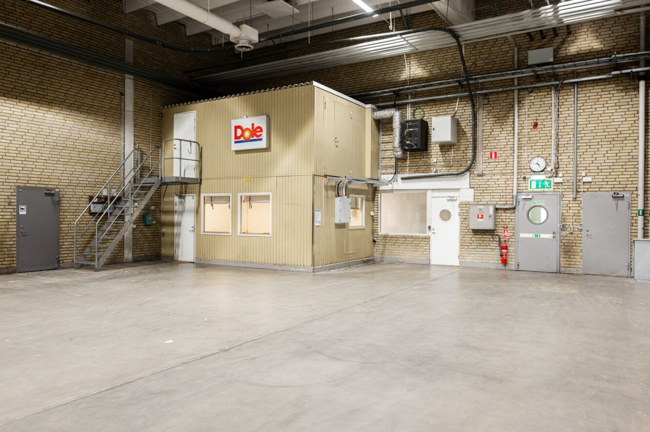 En tom industrilagerlokal med en liten kontorsbyggnad märkt Dole inuti, metalltrappor, tegelväggar och synliga rör och dörrar.