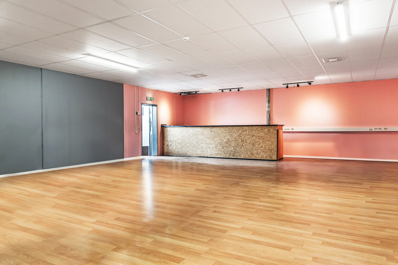 Stort tomt rum med ljust trägolv, en disk med en front av träpanel, laxrosa och grå väggar och lysrör i taket.