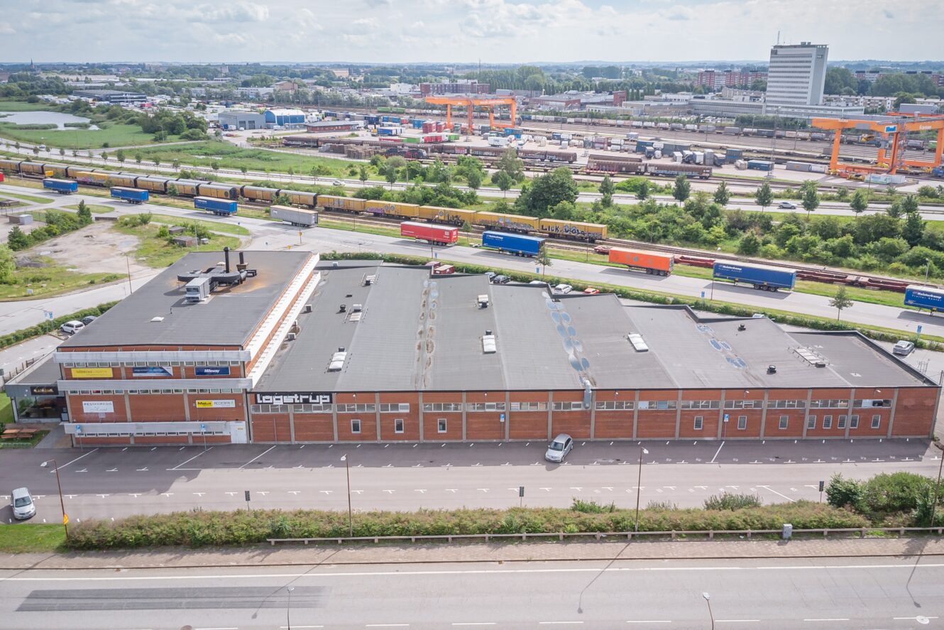 Flygfoto över en stor lagerlokal med Logentryp-skyltar, parkerade lastbilar i närheten, en rad fraktcontainrar och järnvägsspår i ett industriområde.