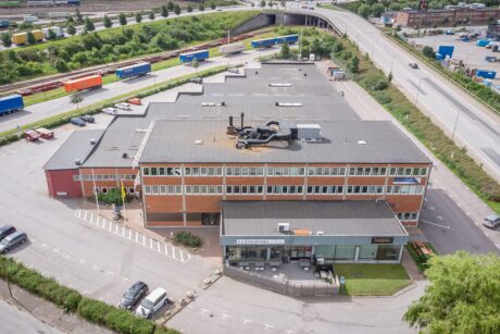 Flygfoto över en stor industri- eller handelsbyggnad med parkeringsplats, omgiven av vägar, lastbilar och grönska.