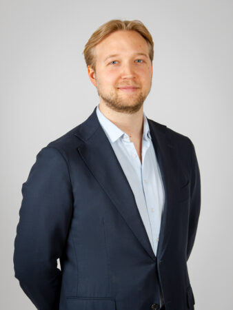 En man med ljusbrunt hår och kort skägg, klädd i en marinblå kostymjacka över en ljusblå skjorta, står mot en ljusgrå bakgrund, vänd mot kameran och med ett lätt leende på läpparna.