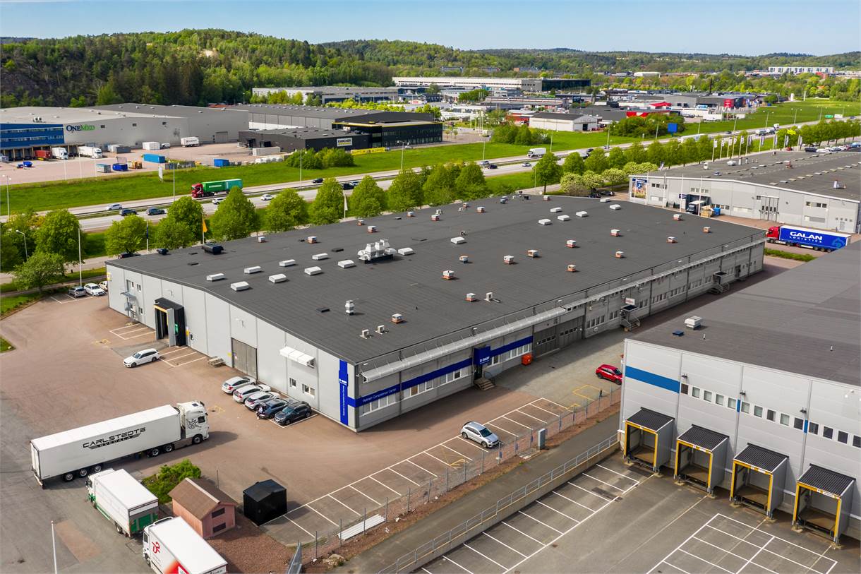 Flygfoto över en stor industrihall med mörkt tak, lastkajer, parkerade lastbilar och närliggande vägar, omgiven av grönska och andra kommersiella byggnader.