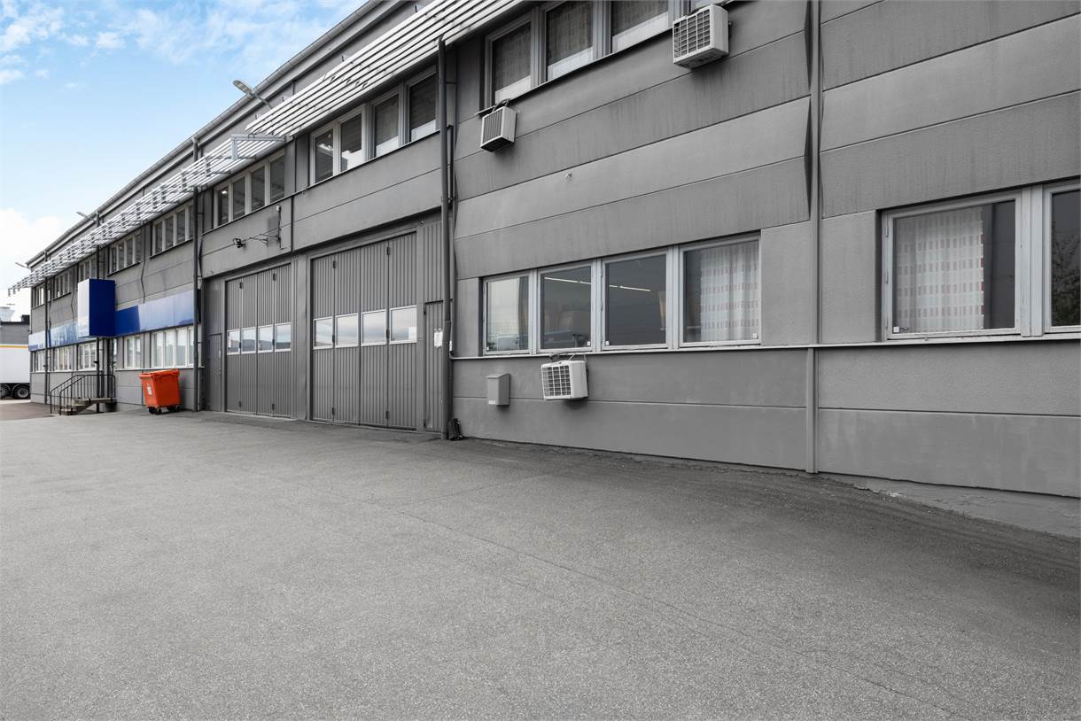 Grå industribyggnad med stora garageportar, flera fönster, luftkonditioneringsaggregat och en röd container på en tom asfalterad tomt.