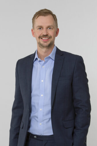 En man i blå kostym och ljusblå skjorta står och ler mot en ljusgrå bakgrund.