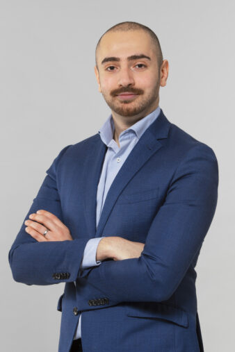 En man i blå kostym och ljusblå skjorta står med armarna i kors mot en ljusgrå bakgrund.