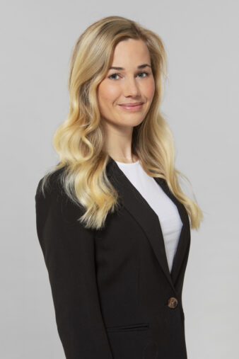 En kvinna med långt blont hår, klädd i svart kavaj och vit topp, står mot en ljusgrå bakgrund.