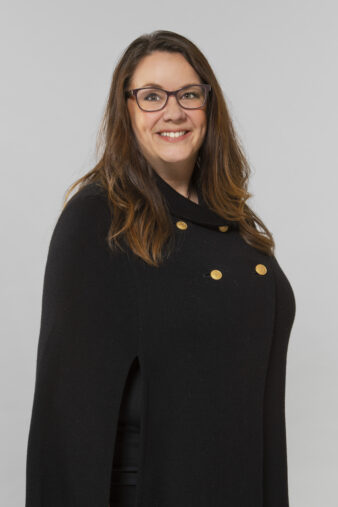 En kvinna med långt brunt hår och glasögon, klädd i en svart kappa med guldknappar, står vänd lite åt sidan och ler mot kameran mot en enfärgad bakgrund.