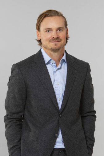En man med ljusbrunt hår, klädd i mörkgrå kostym och ljusblå skjorta, står mot en ljusgrå bakgrund, vänd mot kameran och med ett lätt leende på läpparna.