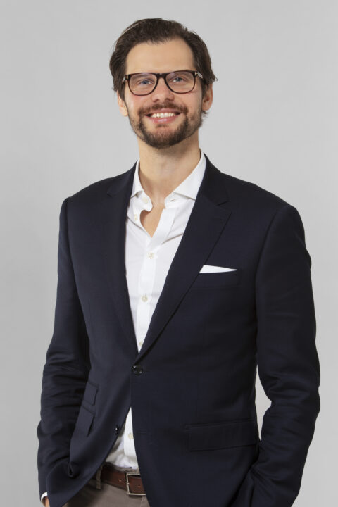 En man med glasögon och skägg, klädd i mörk kostymjacka och vit skjorta, står mot en ljusgrå bakgrund och ler.