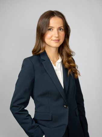 En kvinna med långt brunt hår, klädd i mörk kostymjacka och vit skjorta, står mot en ljus bakgrund och tittar in i kameran.