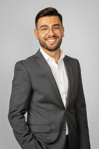 En man med glasögon och grå kostym med vit skjorta står mot en ljusgrå bakgrund och ler mot kameran.