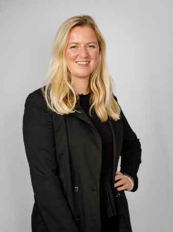 En kvinna med långt blont hår och svart kavaj står mot en ljusgrå bakgrund och ler mot kameran med ena handen på höften.