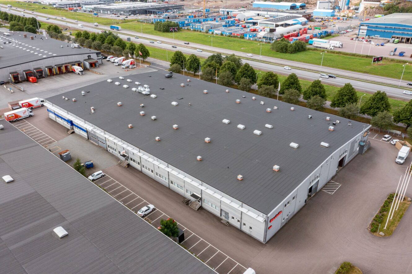 Flygfoto över en stor industrihall med platt tak, omgiven av parkeringsplatser och belägen nära en motorväg med fordon och träd längs vägen.