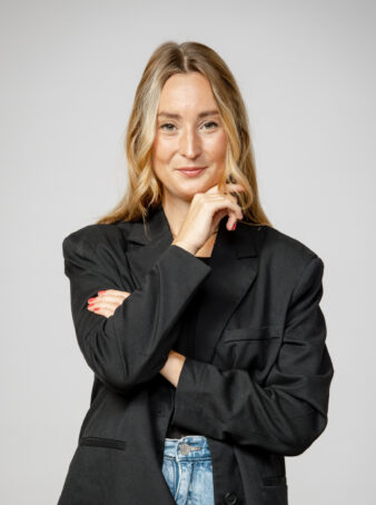 En kvinna med långt blont hår, svart kavaj och jeans står med ena armen i kors och handen vilande på hakan och poserar mot en ljusgrå bakgrund.