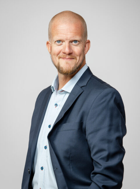 En skallig man med skägg, klädd i blå kostymjacka och ljusblå skjorta, står och ler mot en ljus bakgrund.