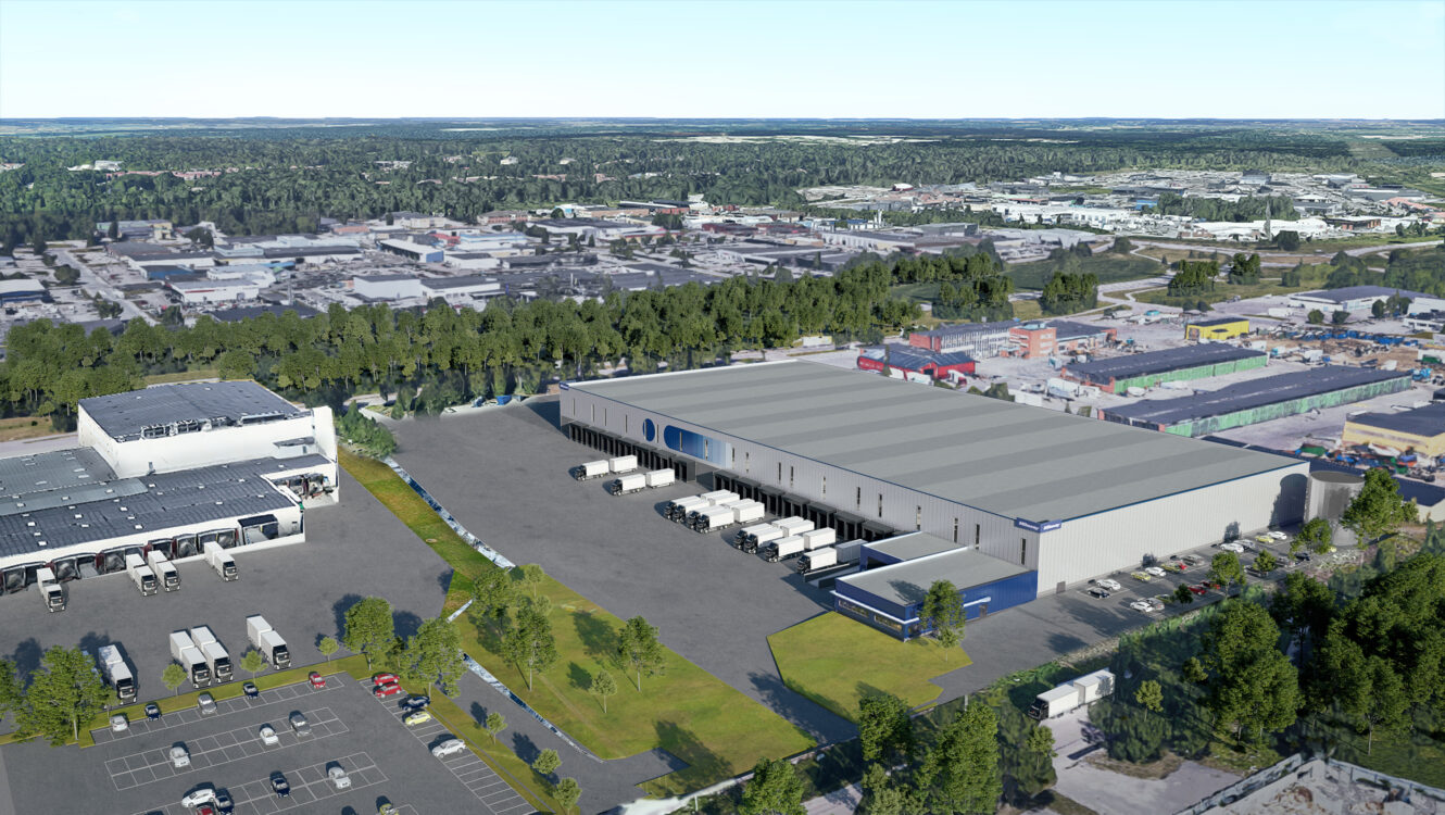 En stor lagerlokal med flera lastkajer och parkerade lastbilar, omgiven av träd och andra industribyggnader i ett stadsområde.