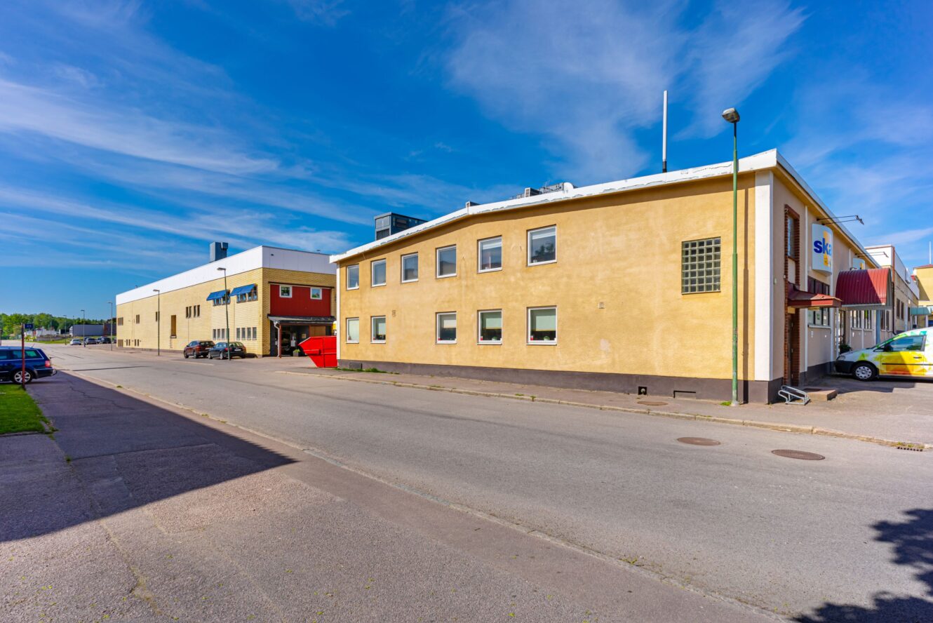 En lugn gata med gula och beigefärgade affärshus under en klarblå himmel; en röd sopcontainer och en parkerad gul bil syns.