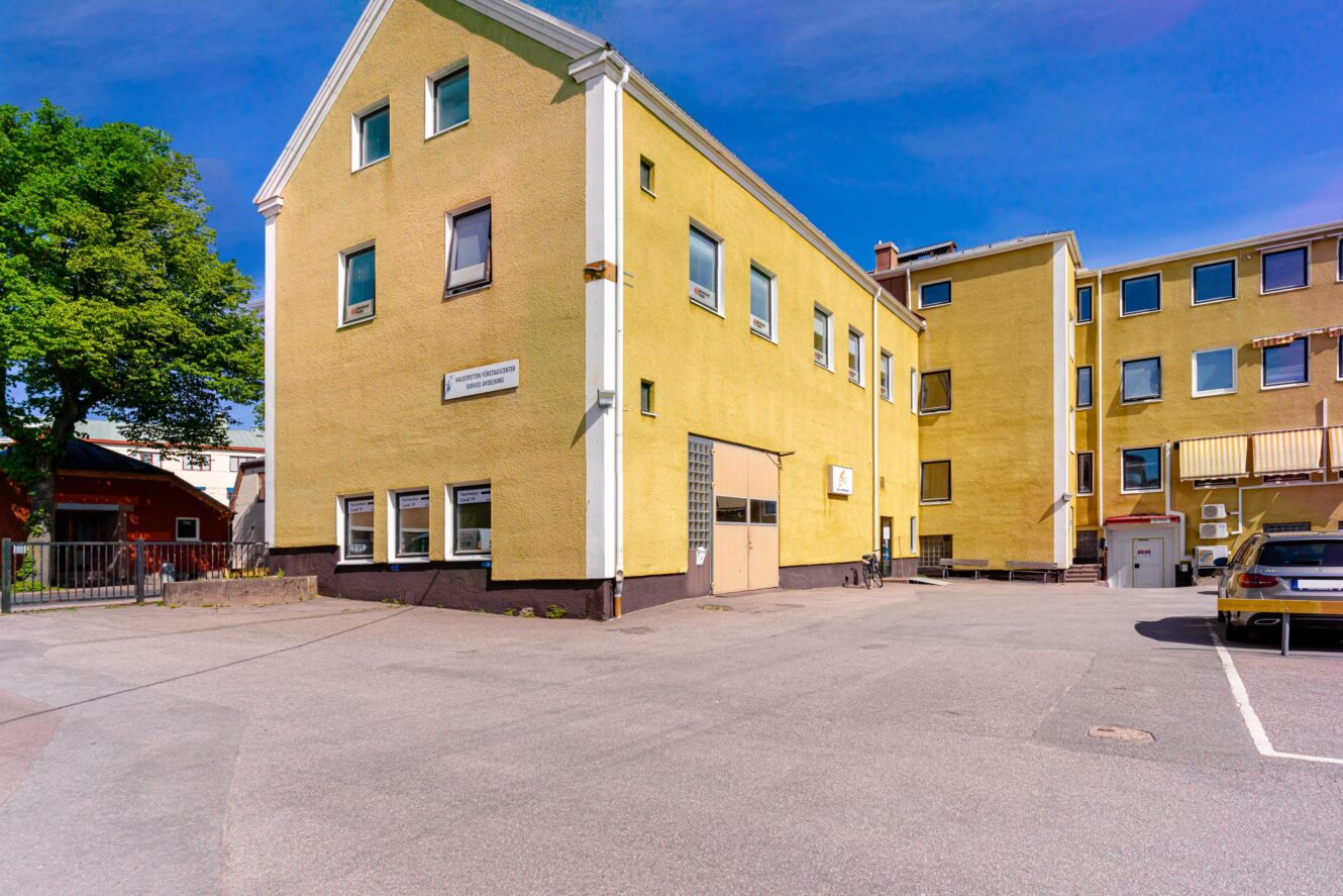 En gul trevåningsbyggnad med flera fönster, en central garageport och en asfalterad parkeringsplats med en bil parkerad till höger.