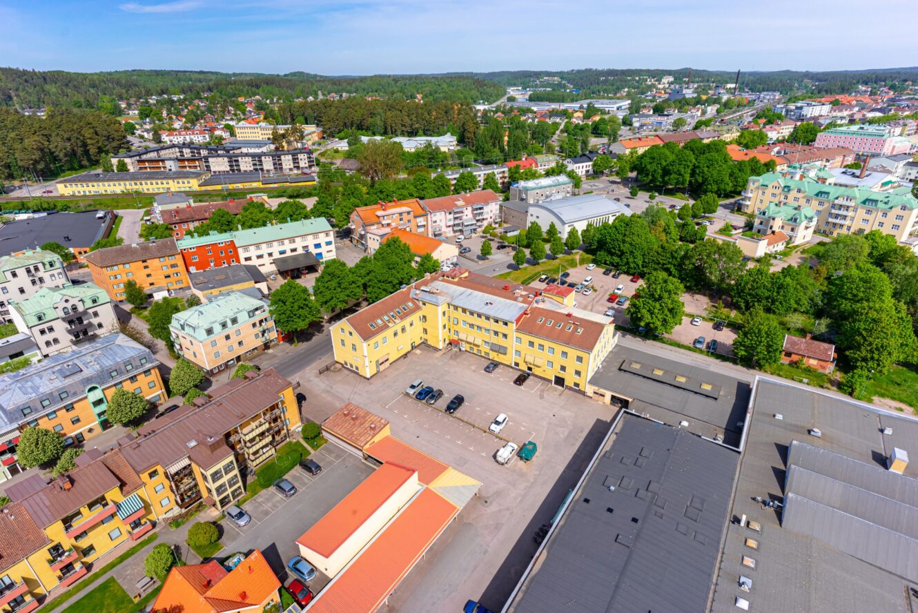 Flygfoto över en förortsstad med bostadshus, gröna träd, parkeringsplatser och industritak under en klar himmel.