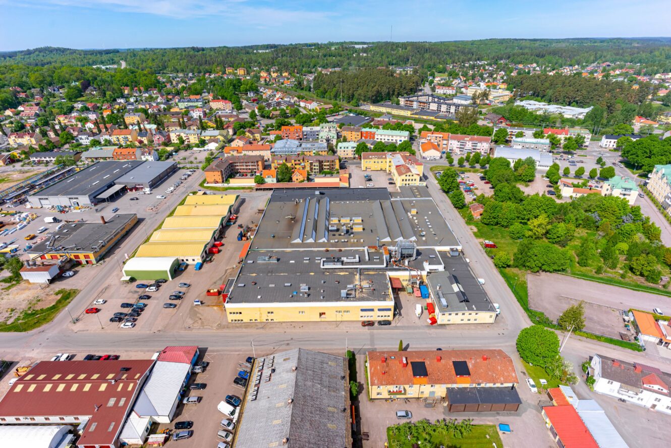 Flygfoto över ett industrikomplex omgivet av kommersiella byggnader, hus, parkerade bilar och träd i ett förortsområde.