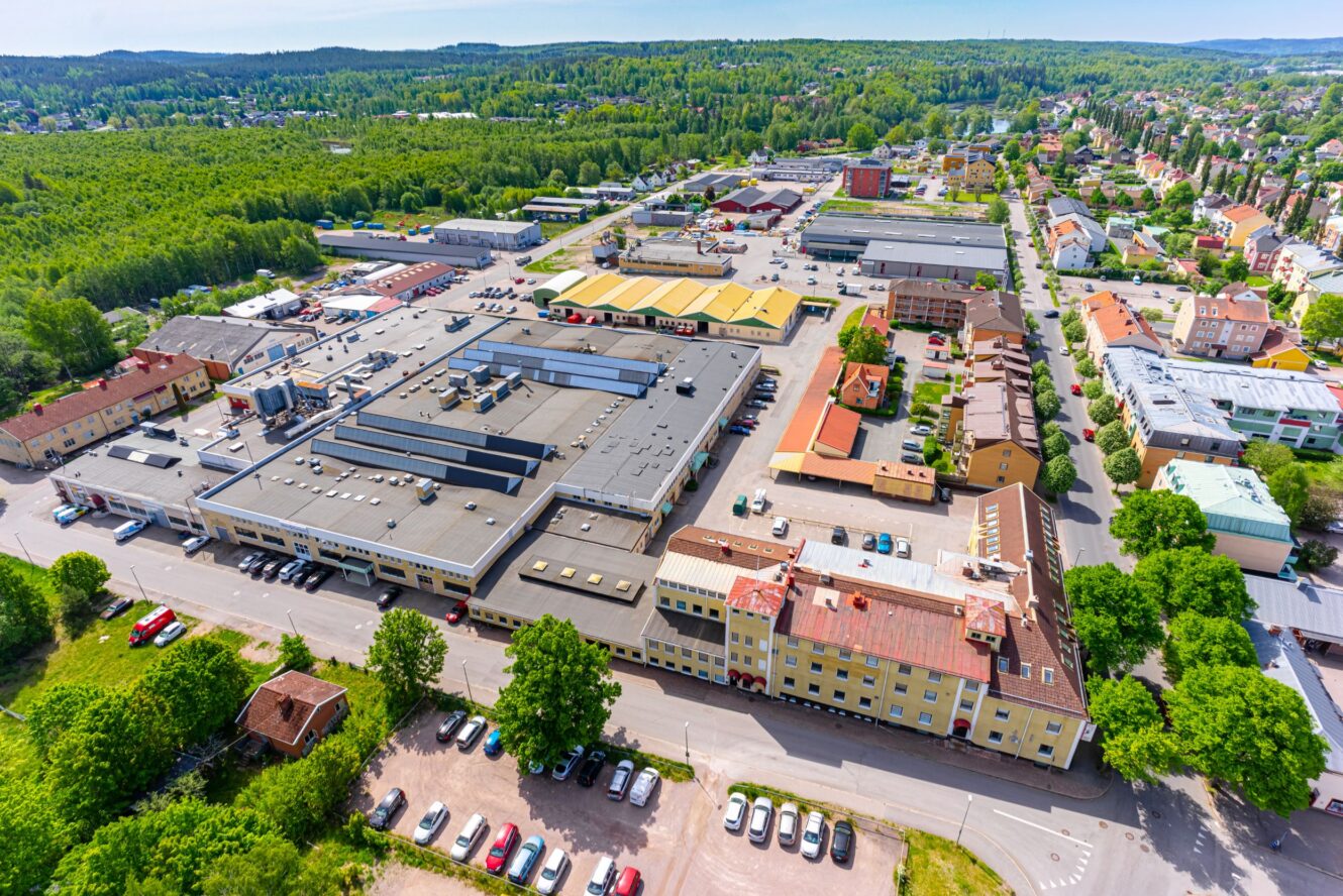 Flygbild över ett industriområde med lagerlokaler, parkeringsplatser och intilliggande bostadshus omgivna av grönska och träd.