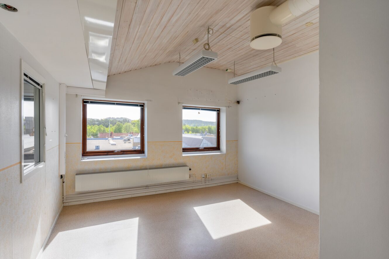 Tomt rum med två stora fönster, vita väggar, trätak, taklampor, element och utsikt över träd och byggnader utanför.