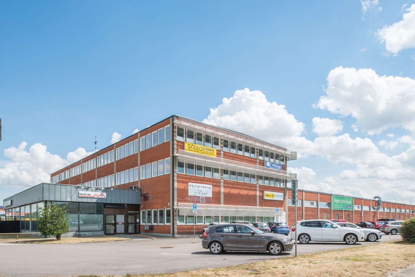En trevånings affärsbyggnad i tegel med glasfönster, flera företagsskyltar, en parkeringsplats och parkerade bilar under en delvis molnig himmel.
