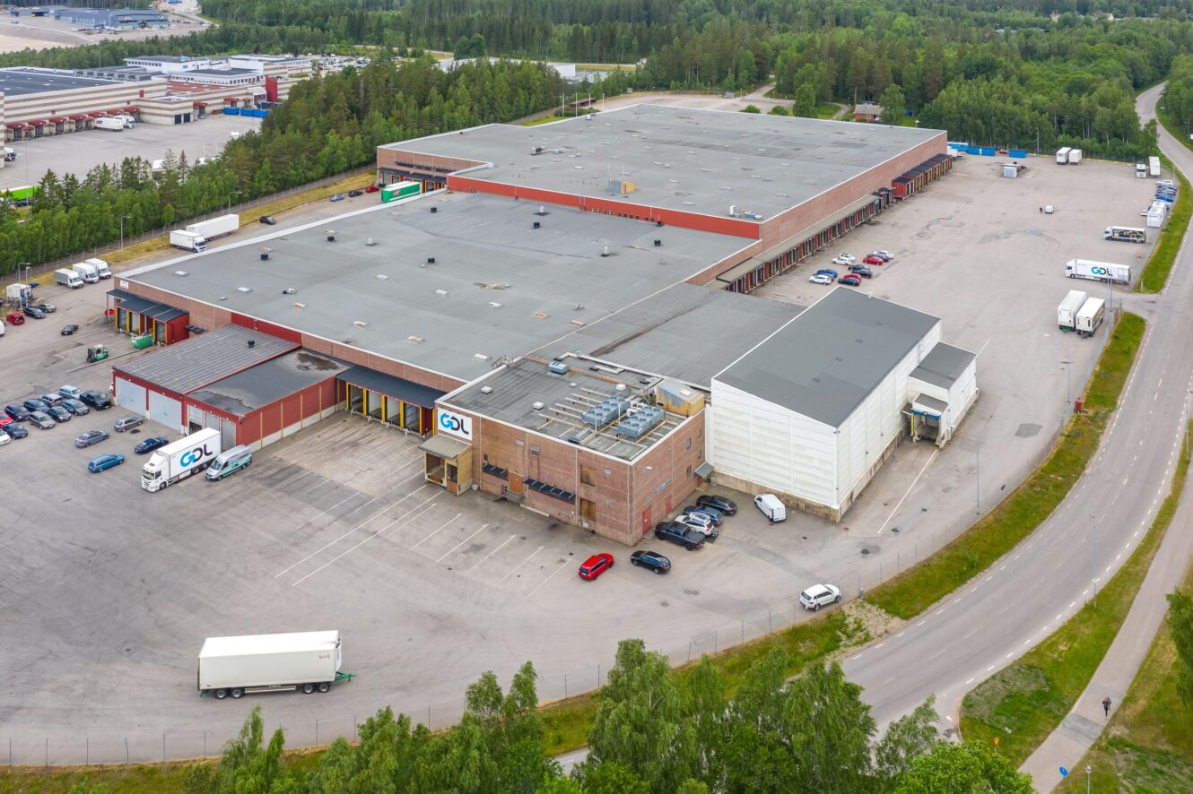 Flygfoto över en stor lagerlokal eller distributionscentral med flera lastkajer, parkerade lastbilar och bilar, omgiven av träd och vägar.