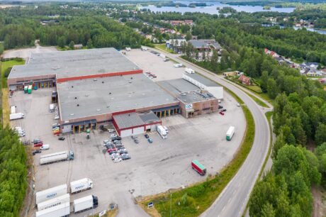 Flygfoto över en stor lagerbyggnad med lastkajer, parkerade lastbilar och bilar, omgiven av träd och vägar i ett lantligt område.