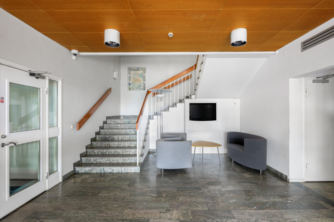 Modern lobby med grått stengolv, två gråa soffor, ett runt bord, en väggmonterad TV, en trappa med ledstång i trä och en vit dörr till vänster.