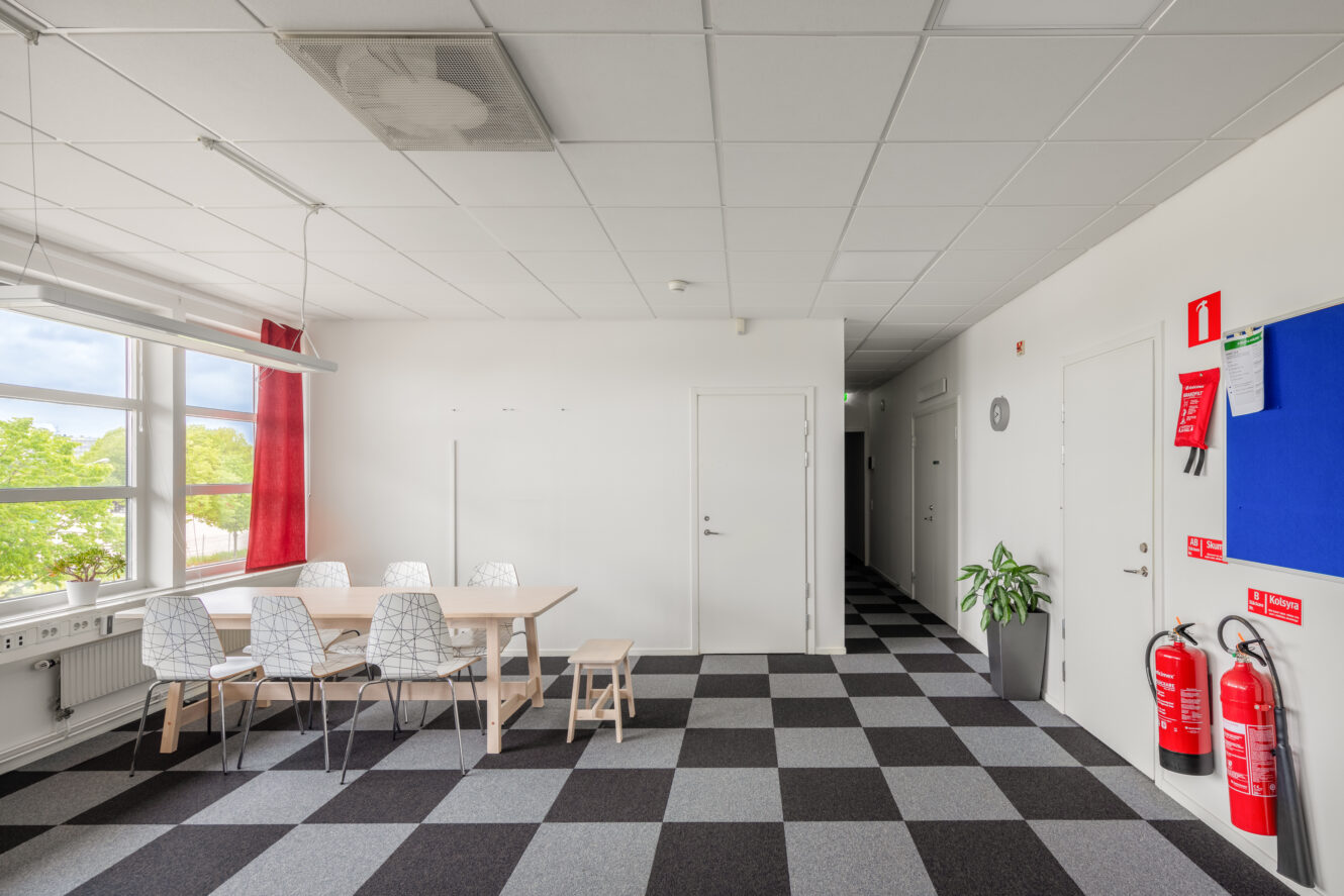 Ett ljust fikarum med träbord, vita stolar, en bänk, stora fönster och rutig matta; brandsläckare och anslagstavla på höger vägg.