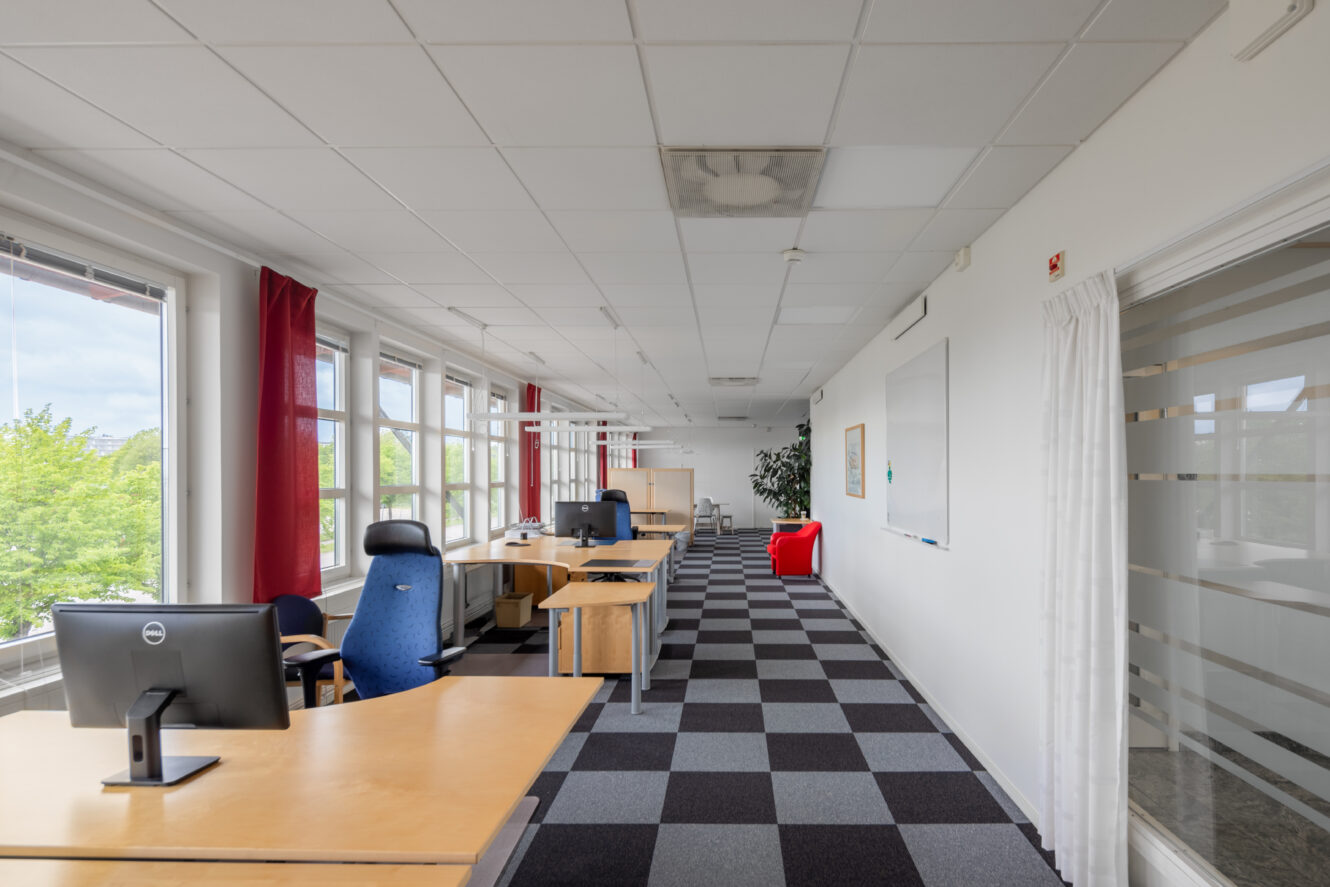 Modern kontorslokal med stora fönster, flera skrivbord och stolar, en datorskärm, en whiteboard och en röd soffa på en rutig heltäckningsmatta.