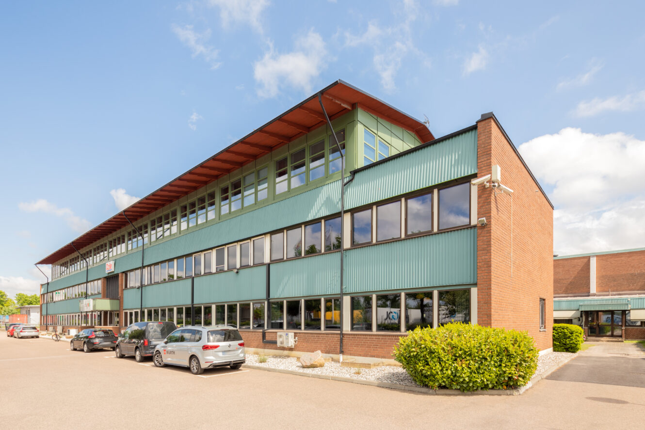 En modern kontorsbyggnad i tre våningar med stora fönster, tegel och grön panel, och flera parkerade bilar längs framsidan, under en delvis molnig himmel.