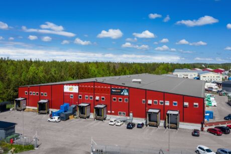 En stor röd lagerbyggnad med flera lastbryggor och parkerade fordon, omgiven av träd och andra industribyggnader under en blå himmel med spridda moln.