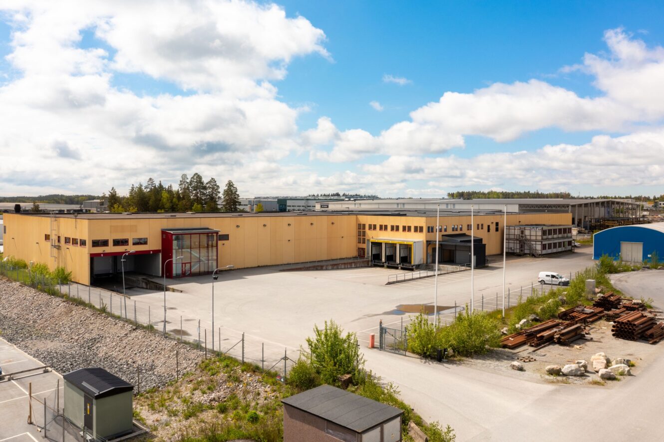 Stort industrilager med lastkajer och stängsel, omgivet av öppen mark, utspridda material och grönska under en delvis molnig himmel.