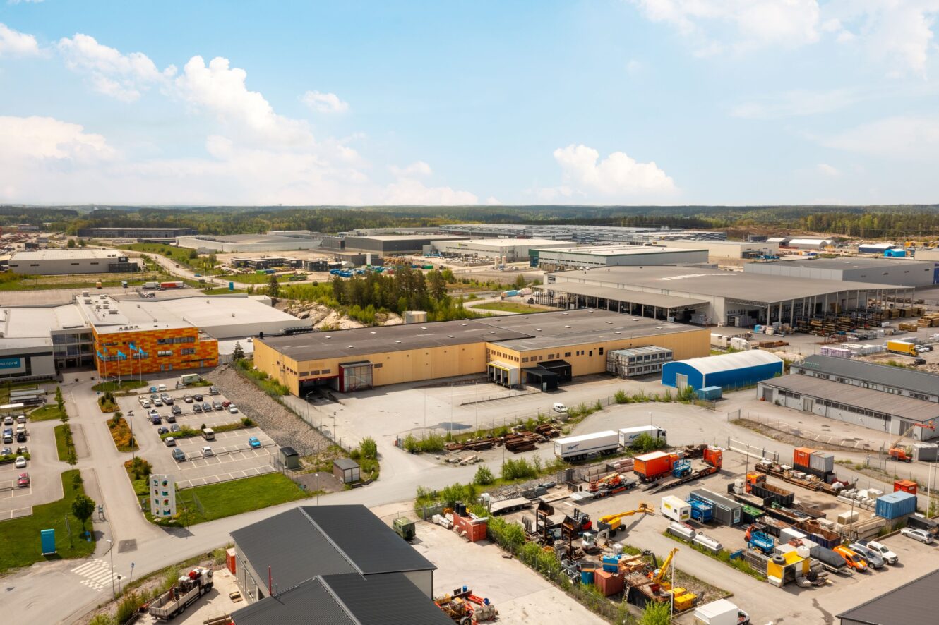 Flygfoto över en industripark med lagerbyggnader, upplag, parkerade lastbilar och diverse utrustning under en delvis molnig himmel.