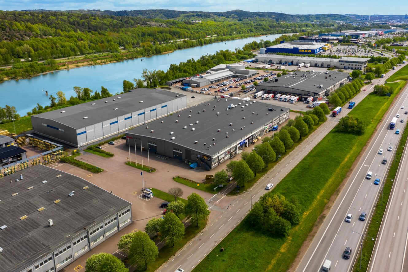 Flygfoto över en industripark med lagerbyggnader, parkeringsplatser, en flod till vänster och en motorväg med bilar till höger, omgiven av gröna träd och kullar.