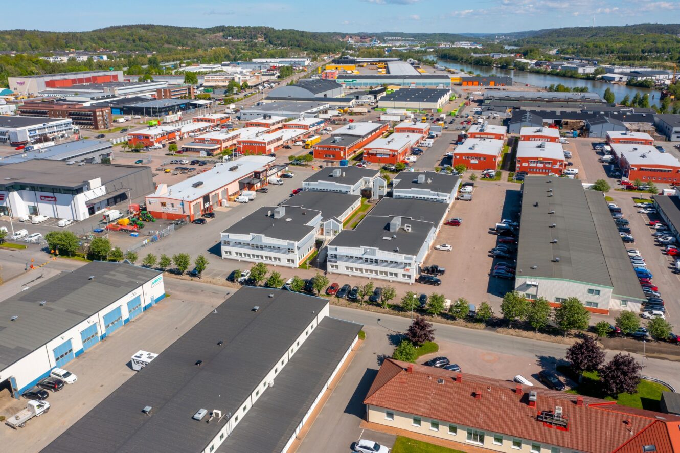 Flygfoto över en industripark med lagerlokaler, kontorsbyggnader, parkeringsplatser och vägar, omgiven av grönska och en vattenväg i bakgrunden.